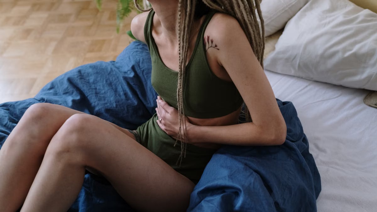 Woman sitting in bed holding her stomach, indicating pain or discomfort, possibly due to cramps.