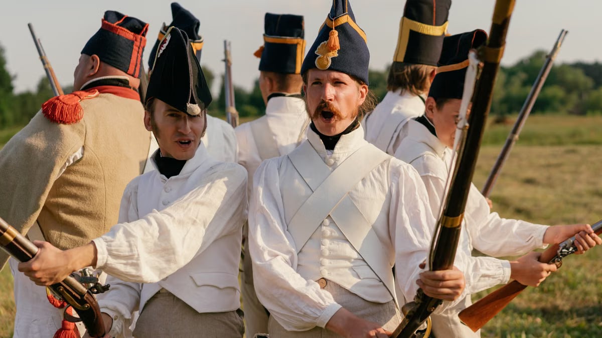 Men in historical military uniforms participate in a reenactment outdoors.